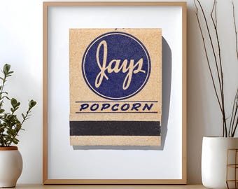 May include: A framed print featuring a vintage-style matchbook cover with a tan background. The design includes a dark blue circle containing the word "jays" in script, with "POPCORN" printed below in blue, and a black strike strip at the bottom.