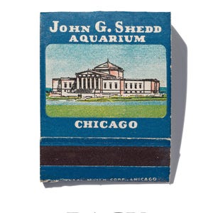 Peut inclure: Une bo&icirc;te d'allumettes vintage illustrant en bleu et blanc l'aquarium John G. Shedd &agrave; Chicago. Le texte "John G. Shedd Aquarium" et "Chicago" est imprim&eacute; en blanc sur la bo&icirc;te d'allumettes.
