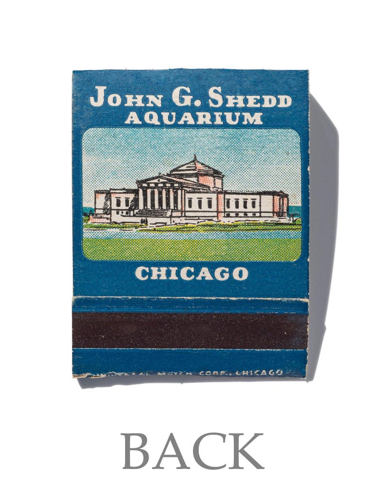 Peut inclure: Une bo&icirc;te d'allumettes vintage illustrant en bleu et blanc l'aquarium John G. Shedd &agrave; Chicago. Le texte "John G. Shedd Aquarium" et "Chicago" est imprim&eacute; en blanc sur la bo&icirc;te d'allumettes.