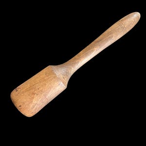 May include: A wooden mallet with a cylindrical head and a long, tapered handle. The wood features a natural light brown grain with visible texture, minor surface scratches, and two thin, circular engraved lines near the end of the handle.