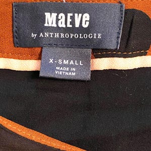 以下が含まれることがあります： 「Maeve by Anthropologie」のブランド名が記載された衣類のタグのクローズアップ。タグはネイビーブルーで、白い文字でX-Smallサイズと表示されています。衣類は黒と茶色の模様です。