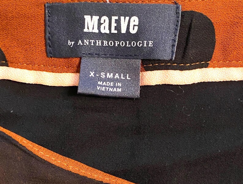 以下が含まれることがあります： 「Maeve by Anthropologie」のブランド名が記載された衣類のタグのクローズアップ。タグはネイビーブルーで、白い文字でX-Smallサイズと表示されています。衣類は黒と茶色の模様です。