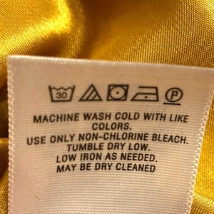 Op de afbeelding: Close-up van een wit stoffen label met wasvoorschriften tegen een gele satijnen achtergrond. Het label bevat symbolen voor wassen op 30 graden, bleken, drogen in de droger, strijken en chemisch reinigen. Tekst op het label geeft gedetailleerde wasinstructies.