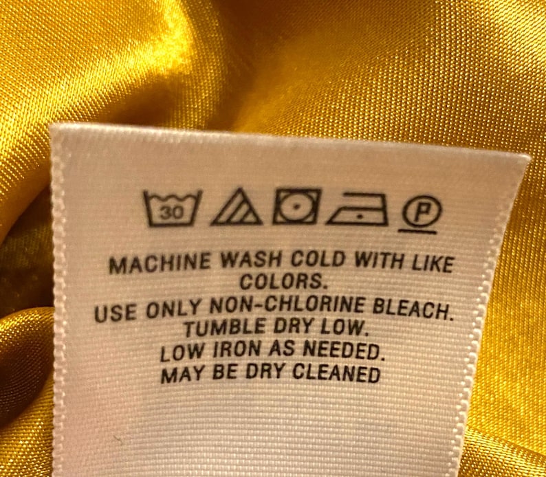 Op de afbeelding: Close-up van een wit stoffen label met wasvoorschriften tegen een gele satijnen achtergrond. Het label bevat symbolen voor wassen op 30 graden, bleken, drogen in de droger, strijken en chemisch reinigen. Tekst op het label geeft gedetailleerde wasinstructies.