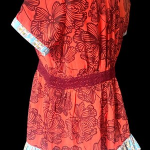 May include: A red floral print top with short sleeves, a burgundy lace waist, and a ruffled hem. The sleeves and hem feature a blue and white patterned trim. The top has a V-neck and a loose fit.