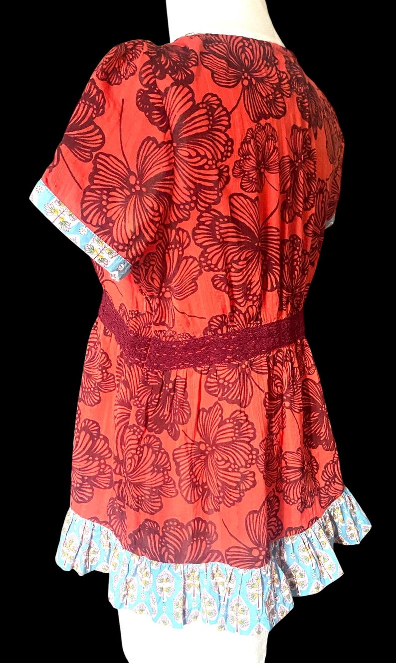 May include: A red floral print top with short sleeves, a burgundy lace waist, and a ruffled hem. The sleeves and hem feature a blue and white patterned trim. The top has a V-neck and a loose fit.