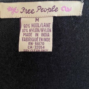 May include: Close-up of a black garment tag with the brand name "Free People" in pink script. The tag also includes size "M", fabric composition, and "Made in India" text. The garment is made of 90% wool and 10% nylon.