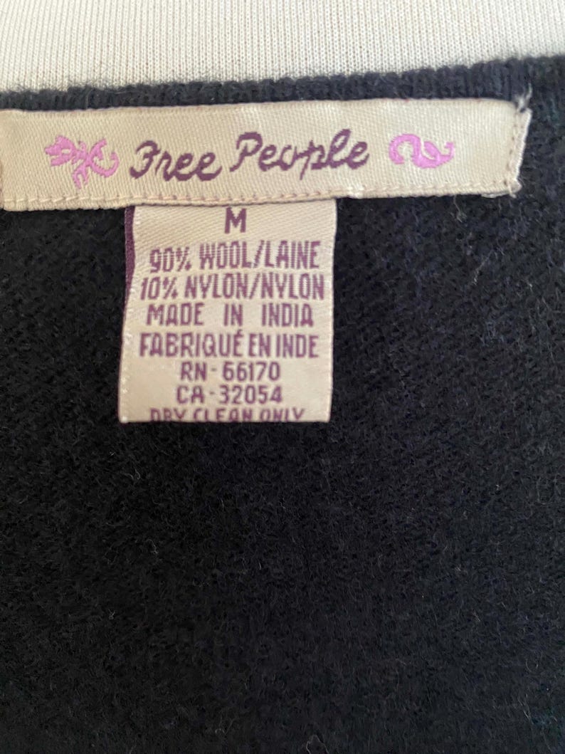 May include: Close-up of a black garment tag with the brand name "Free People" in pink script. The tag also includes size "M", fabric composition, and "Made in India" text. The garment is made of 90% wool and 10% nylon.