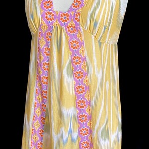 May include: Yellow sleeveless dress with a V-neckline. The dress features a colorful pattern and is trimmed with a purple floral design. The dress is displayed on a mannequin against a black background.