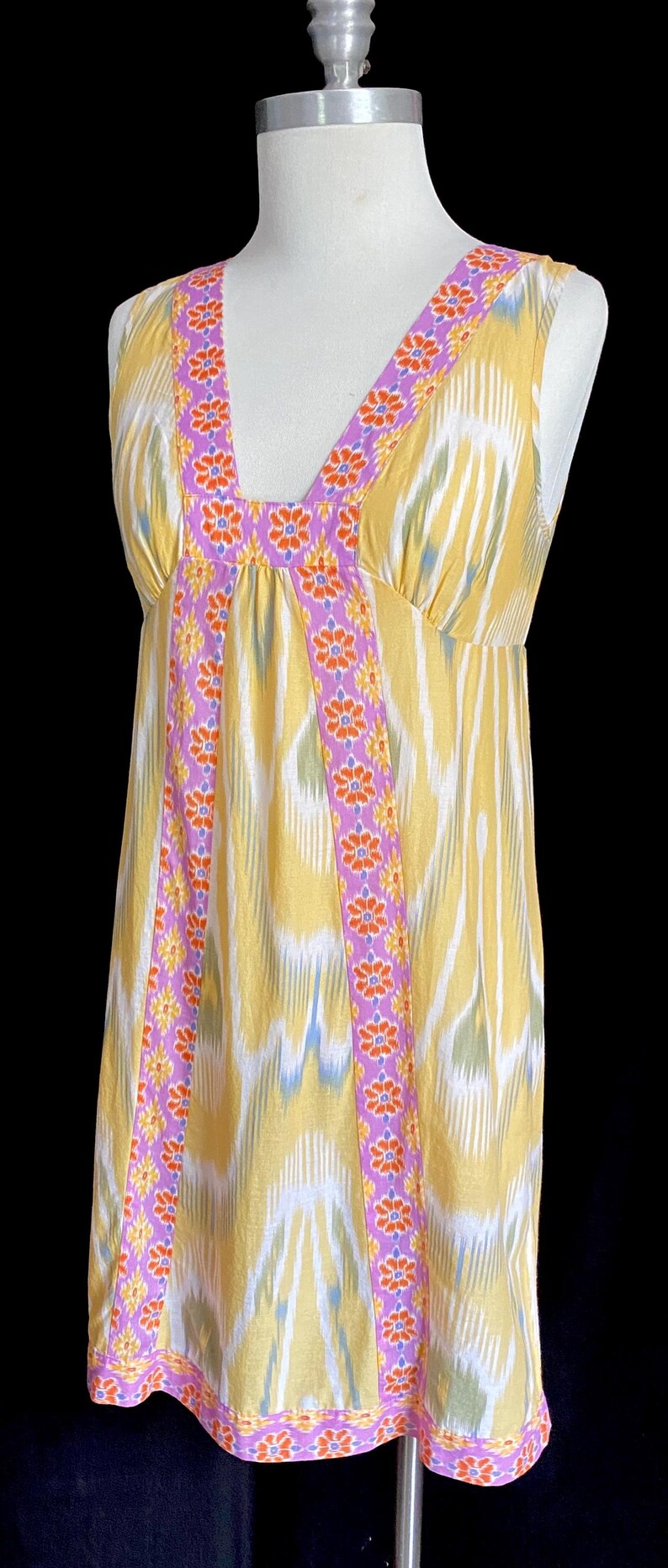 May include: Yellow sleeveless dress with a V-neckline. The dress features a colorful pattern and is trimmed with a purple floral design. The dress is displayed on a mannequin against a black background.