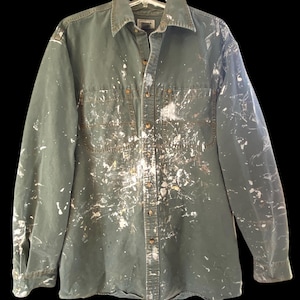 May include: A long-sleeved, olive green button-down shirt heavily splattered with white paint. The shirt has two chest pockets and a collar. The paint is distributed across the front, sleeves, and back of the shirt.
