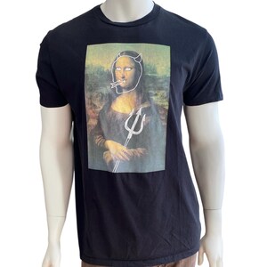 May include: Black t-shirt featuring a parody of the Mona Lisa with devil horns, a cigarette, and a trident. The artwork is a humorous take on the classic painting, with a white outline overlaying the original image. The shirt is displayed on a mannequin.