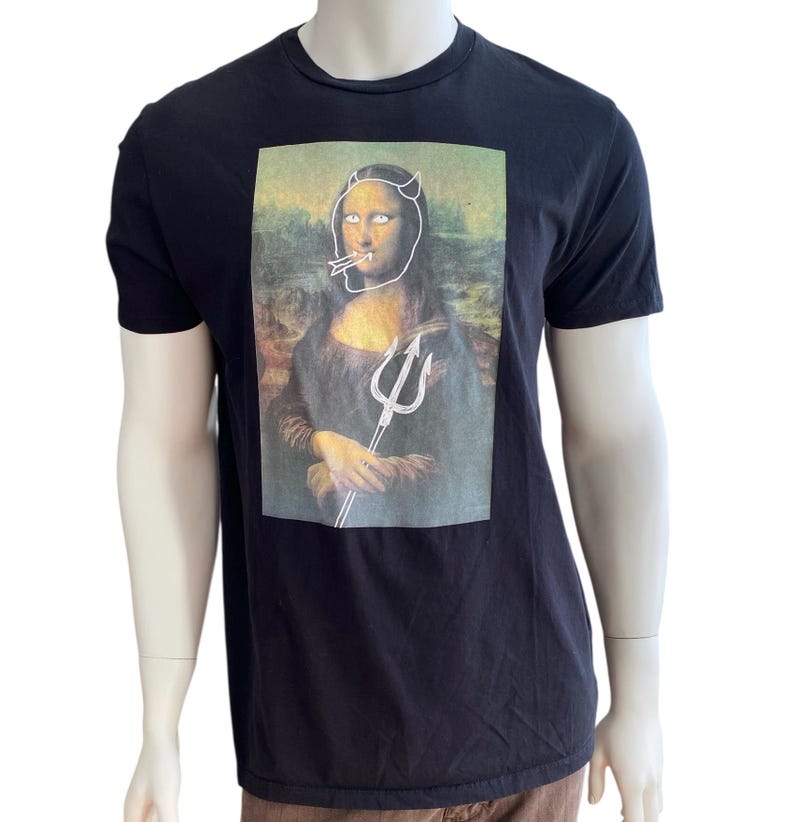 May include: Black t-shirt featuring a parody of the Mona Lisa with devil horns, a cigarette, and a trident. The artwork is a humorous take on the classic painting, with a white outline overlaying the original image. The shirt is displayed on a mannequin.