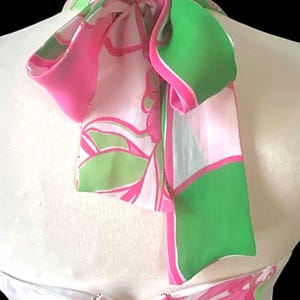 Op de afbeelding: Een witte halterjurk met een levendig roze en groen bloemenpatroon. De jurk heeft een hoge halslijn met een strikdetail en een ritssluiting. De stof heeft een zijdeachtig uiterlijk.