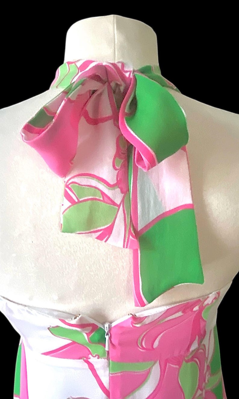 Op de afbeelding: Een witte halterjurk met een levendig roze en groen bloemenpatroon. De jurk heeft een hoge halslijn met een strikdetail en een ritssluiting. De stof heeft een zijdeachtig uiterlijk.