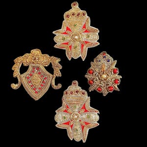 May include: Four decorative embroidered patches featuring gold metallic thread, red fabric accents, and small colorful beads. The designs include two cross-shaped emblems, one shield-shaped piece with ornate scrollwork, and one circular floral motif.