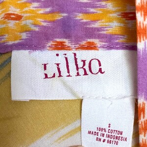 May include: Close-up of a tag from a garment with the brand name "Liika" in red lettering. The tag is white and made of fabric. The garment has a colorful pattern with orange, yellow, and purple hues. The tag also indicates the garment is size S and made of 100% cotton.