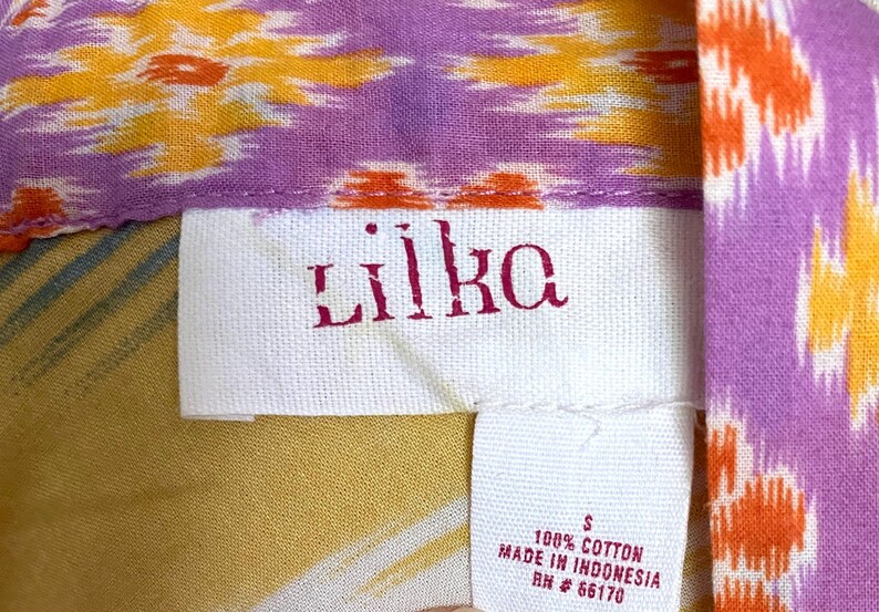 May include: Close-up of a tag from a garment with the brand name "Liika" in red lettering. The tag is white and made of fabric. The garment has a colorful pattern with orange, yellow, and purple hues. The tag also indicates the garment is size S and made of 100% cotton.