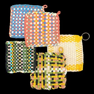 May include: A collection of five square woven potholders featuring various color combinations including blue, white, red, yellow, green, and black. Each potholder has a braided border and a small loop or metal ring attached for hanging purposes.