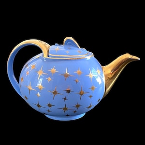 May include: A light blue teapot with a gold handle, spout, and lid. The teapot is decorated with gold starburst patterns. The teapot is a round shape and sits on a black background.