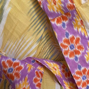 May include: Close-up of a garment featuring a vibrant mix of colors and patterns. The fabric showcases a yellow background with white and blue stripes, complemented by a purple border adorned with orange and yellow floral designs. The stitching is visible.