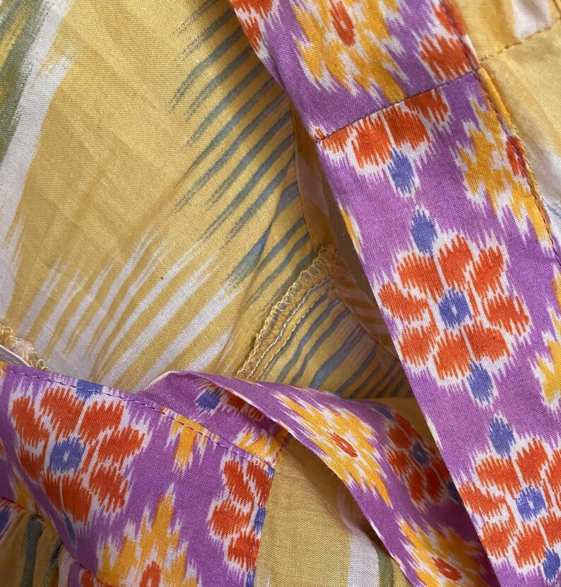 May include: Close-up of a garment featuring a vibrant mix of colors and patterns. The fabric showcases a yellow background with white and blue stripes, complemented by a purple border adorned with orange and yellow floral designs. The stitching is visible.