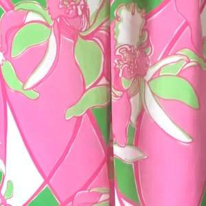 Op de afbeelding: Close-up van een stof met een levendig bloemenpatroon. Het ontwerp toont grote roze bloemen met groene en witte accenten tegen een groen-witte geometrische achtergrond. De algehele esthetiek is helder en kleurrijk.