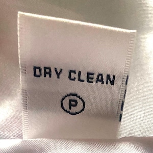 Op de afbeelding: Een wit stoffen label met de woorden "DRY CLEAN" in donkerblauw gedrukt, samen met een cirkel met de letter "P". Dit is een wasvoorschriftlabel.
