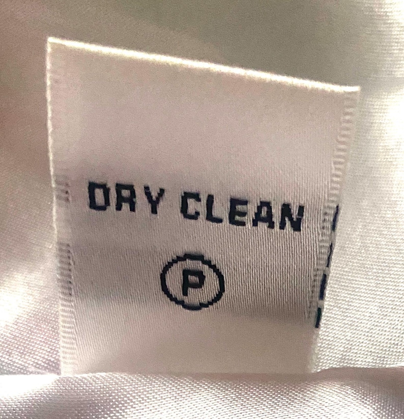 Op de afbeelding: Een wit stoffen label met de woorden "DRY CLEAN" in donkerblauw gedrukt, samen met een cirkel met de letter "P". Dit is een wasvoorschriftlabel.