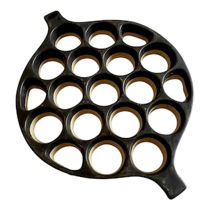 May include: A black cast iron pan with 19 circular indentations, likely for making small pancakes or pastries. The pan has a unique, leaf-like shape with a handle on one end. The interior of each indentation is a light brown color.