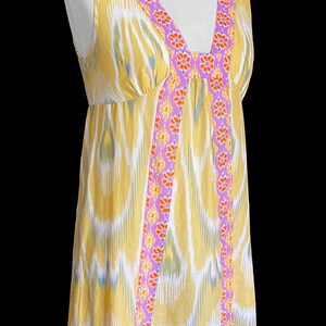 May include: A sleeveless yellow dress with a V-neckline and a flowing silhouette. The dress features a colorful ikat pattern and is trimmed with a purple floral design along the neckline and edges. The dress is displayed on a mannequin.