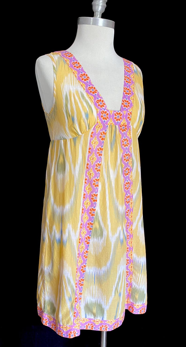 May include: A sleeveless yellow dress with a V-neckline and a flowing silhouette. The dress features a colorful ikat pattern and is trimmed with a purple floral design along the neckline and edges. The dress is displayed on a mannequin.