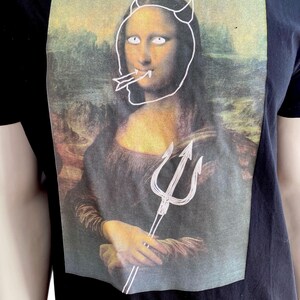 May include: Black t-shirt featuring a graphic of the Mona Lisa with devil horns, a trident, and a tongue sticking out. The artwork is a parody of the famous painting. The shirt is made of a dark fabric.