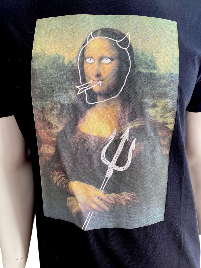 May include: Black t-shirt featuring a graphic of the Mona Lisa with devil horns, a trident, and a tongue sticking out. The artwork is a parody of the famous painting. The shirt is made of a dark fabric.