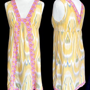 May include: A sleeveless yellow dress with a V-neckline and a colorful floral and geometric pattern. The dress features a pink and orange floral trim along the neckline and down the front. The fabric has a white, blue, and green ikat design.