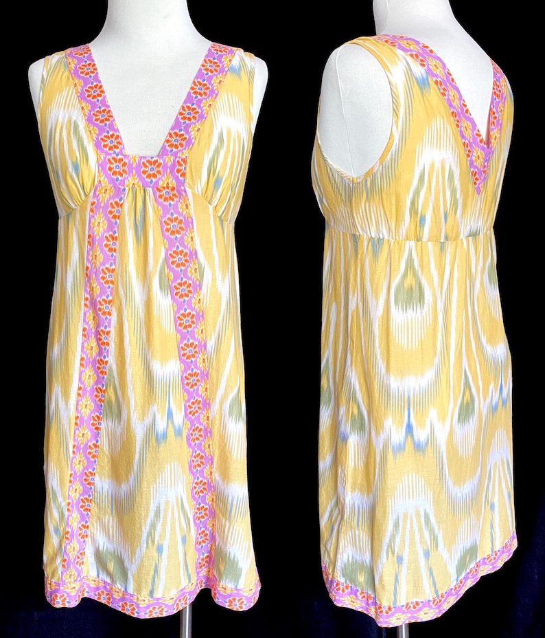 May include: A sleeveless yellow dress with a V-neckline and a colorful floral and geometric pattern. The dress features a pink and orange floral trim along the neckline and down the front. The fabric has a white, blue, and green ikat design.