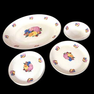 May include: A set of white ceramic dinnerware featuring a central fruit motif of peaches, plums, and berries, with smaller fruit clusters around the rims. The collection includes one large oval platter, two round dinner plates, and two small bowls.