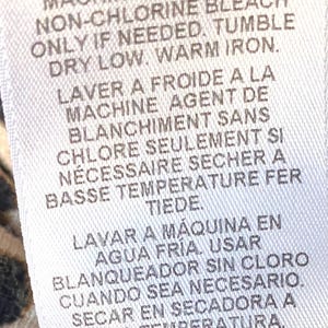May include: White care label with instructions for washing, bleaching, drying, and ironing a garment. The instructions are written in English, French, and Spanish. The label includes symbols for machine wash cold, non-chlorine bleach, tumble dry low, and warm iron.