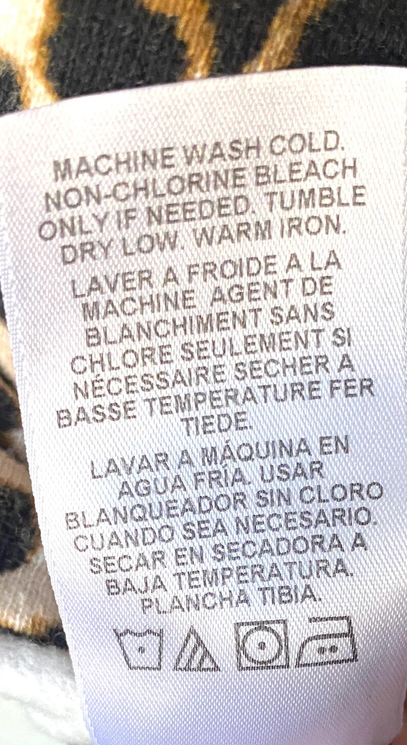 May include: White care label with instructions for washing, bleaching, drying, and ironing a garment. The instructions are written in English, French, and Spanish. The label includes symbols for machine wash cold, non-chlorine bleach, tumble dry low, and warm iron.