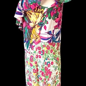 May include: A floral print dress with a white base, featuring large pink, yellow, and blue flowers. The dress has a ruffled neckline and a flowing, floor-length design, perfect for a summer event.