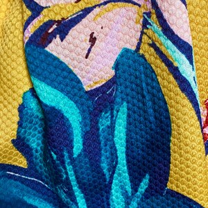 Op de afbeelding: Close-up van een textiel met een bloemenpatroon. Het ontwerp toont grote blauwe en turkooizen bloemen met roze en bordeauxrode accenten tegen een gele achtergrond. De stof heeft een verhoogde, kuiltjesachtige textuur.