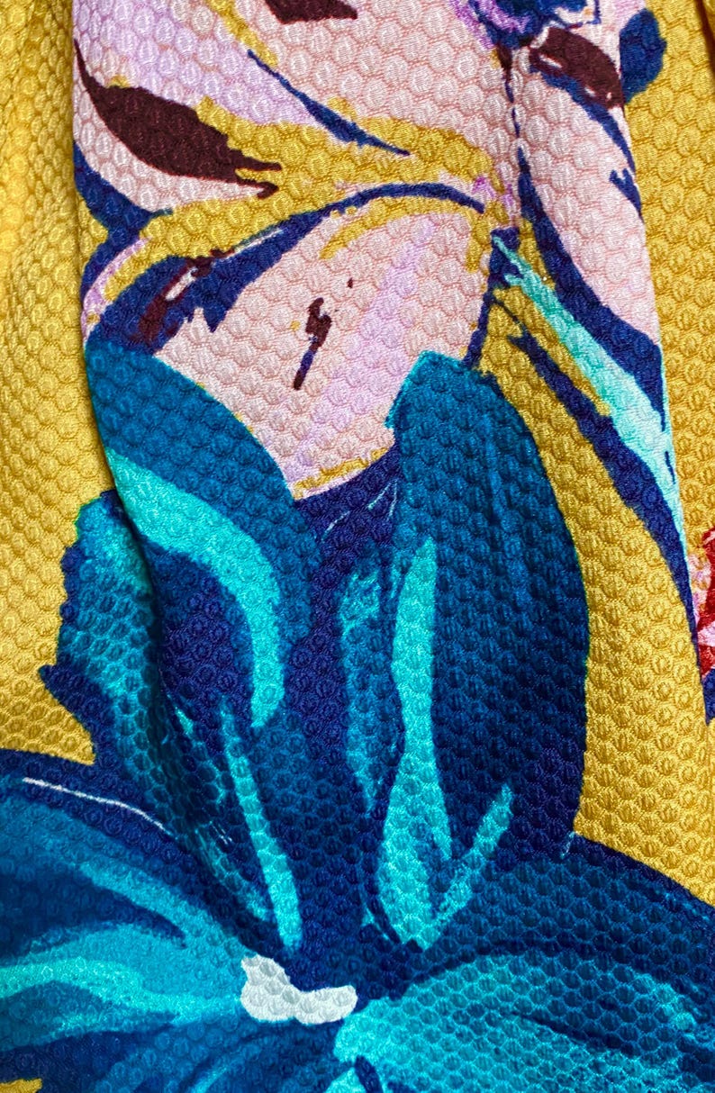 Op de afbeelding: Close-up van een textiel met een bloemenpatroon. Het ontwerp toont grote blauwe en turkooizen bloemen met roze en bordeauxrode accenten tegen een gele achtergrond. De stof heeft een verhoogde, kuiltjesachtige textuur.