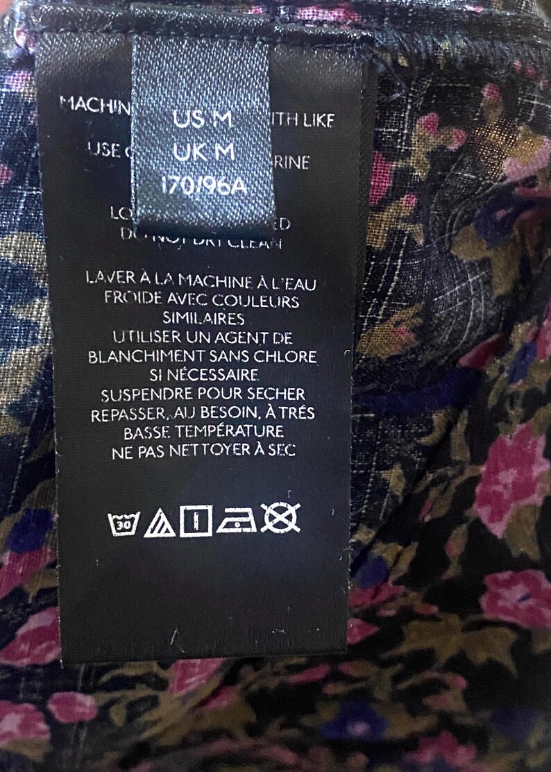 May include: A black clothing tag with white text, including size information: US M, UK M, and 170/96A. The tag also features washing instructions in French, along with care symbols for machine washing at 30 degrees.