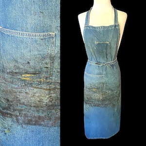 May include: A blue denim apron featuring a bib design with a waist tie and a single front pocket. The fabric displays a weathered appearance with scattered paint splatters in shades of yellow, brown, and dark gray across the lower section.