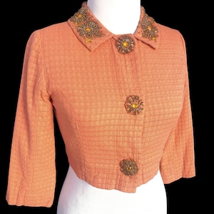 May include: A peach-colored, cropped jacket with a textured weave. The jacket features three-quarter sleeves, a collared neckline, and decorative, flower-shaped buttons. The collar is embellished with intricate beadwork. The jacket is displayed on a white mannequin.