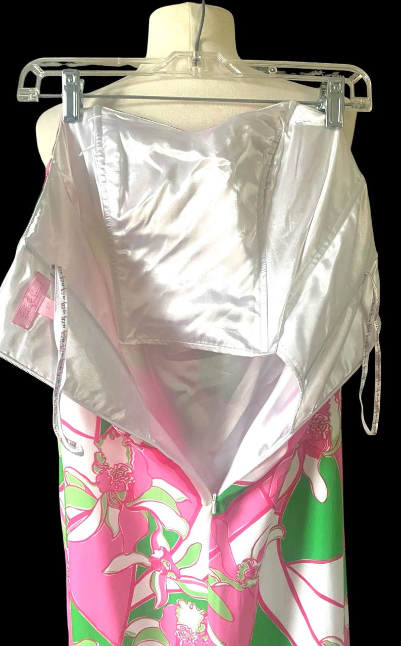 Op de afbeelding: Een strapless jurk met een levendig bloemenpatroon in roze, groen en wit, hangend aan een hanger. De jurk heeft een witte satijnen voering en een ritssluiting. De jurk wordt gepresenteerd tegen een zwarte achtergrond.