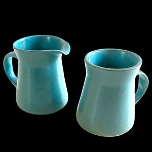 May include: Two turquoise ceramic pitchers with handles. One pitcher has a spout, the other has a rounded rim. Both have a glossy finish and are set against a black background. These pitchers are suitable for serving beverages.