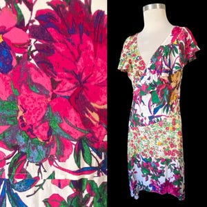 May include: A floral print dress with a V-neck and short sleeves. The dress features a vibrant pattern of pink, green, blue, and yellow flowers on a white background. The dress is displayed on a mannequin against a black backdrop.