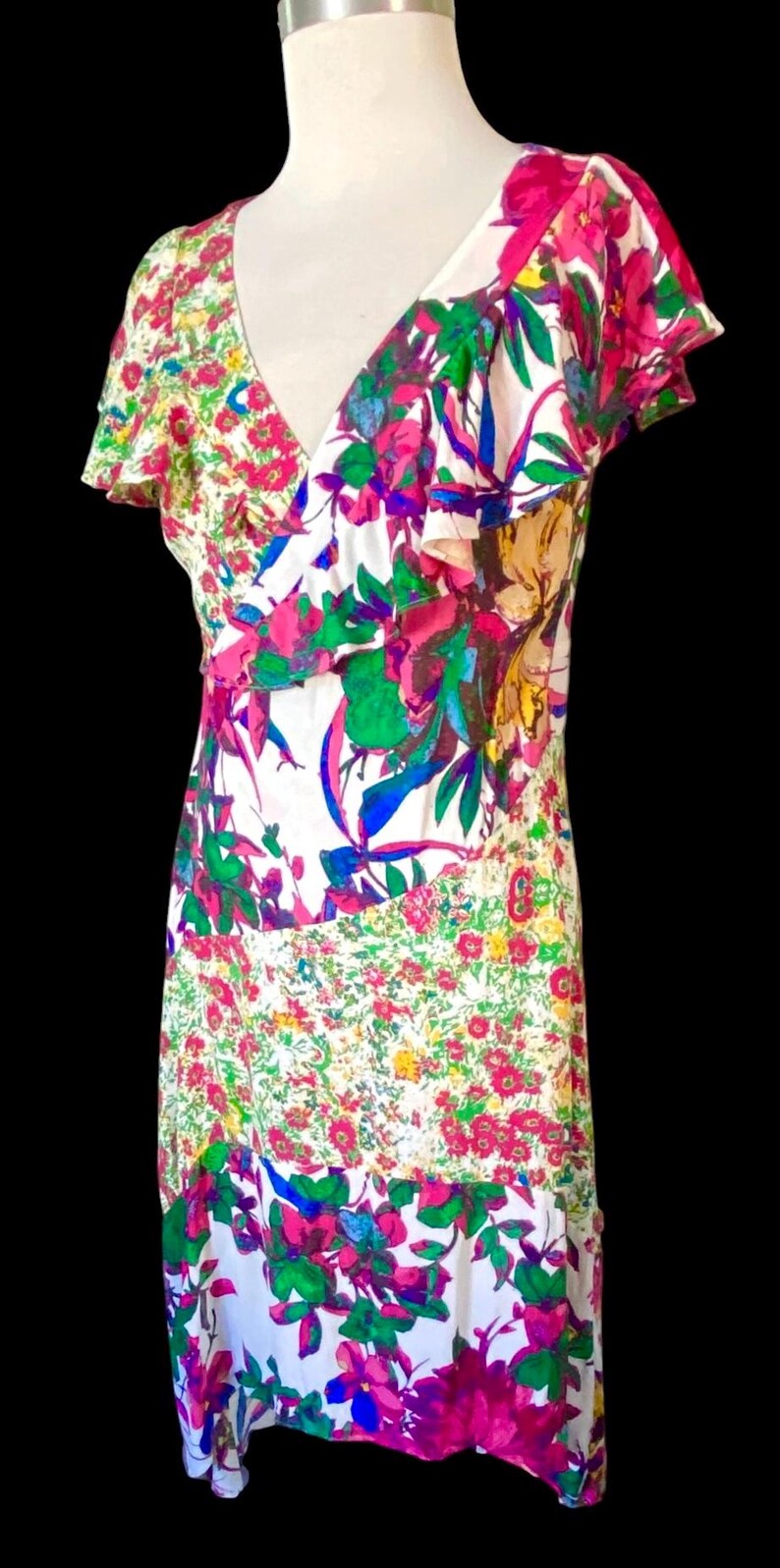 May include: A white, floral-print dress with a V-neckline and ruffled sleeves. The dress features a patchwork design with pink, green, blue, and yellow flowers. The dress is sleeveless and has a flowing skirt.