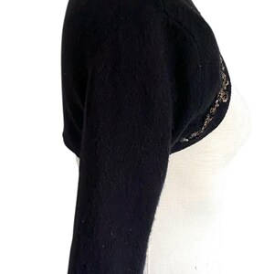 May include: Black bolero sweater with three-quarter sleeves. The sweater has a rounded neckline and is adorned with a decorative, beaded trim along the sleeve cuffs and the front opening. The garment is displayed on a white mannequin.